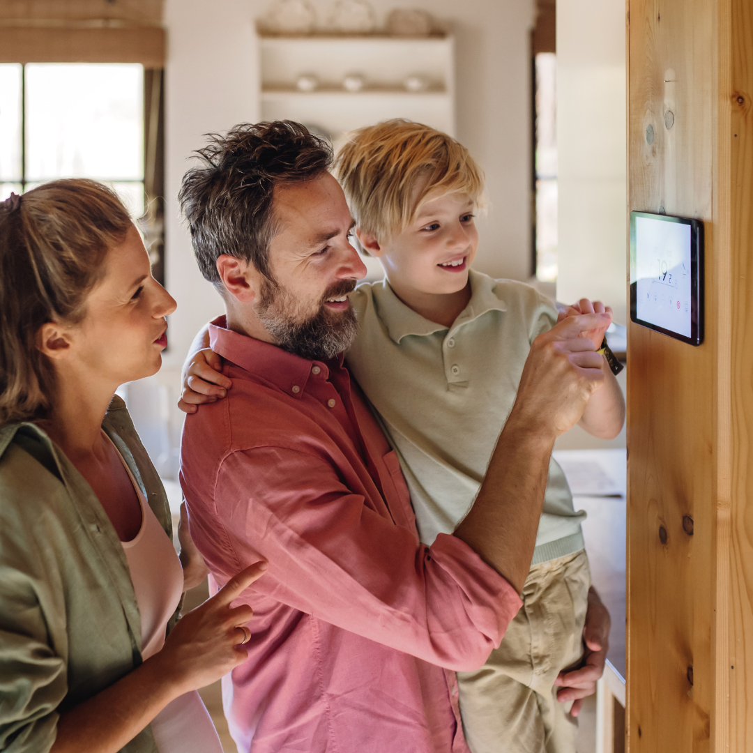 Family using a smart home device on a wall: father holds child, mother points. Light interior.