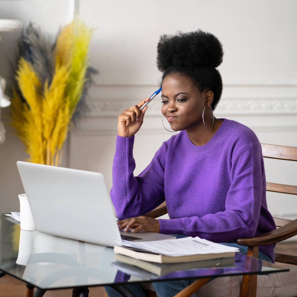 Woman in purple sweater, seated at desk with laptop, holding pen, looking thoughtful.