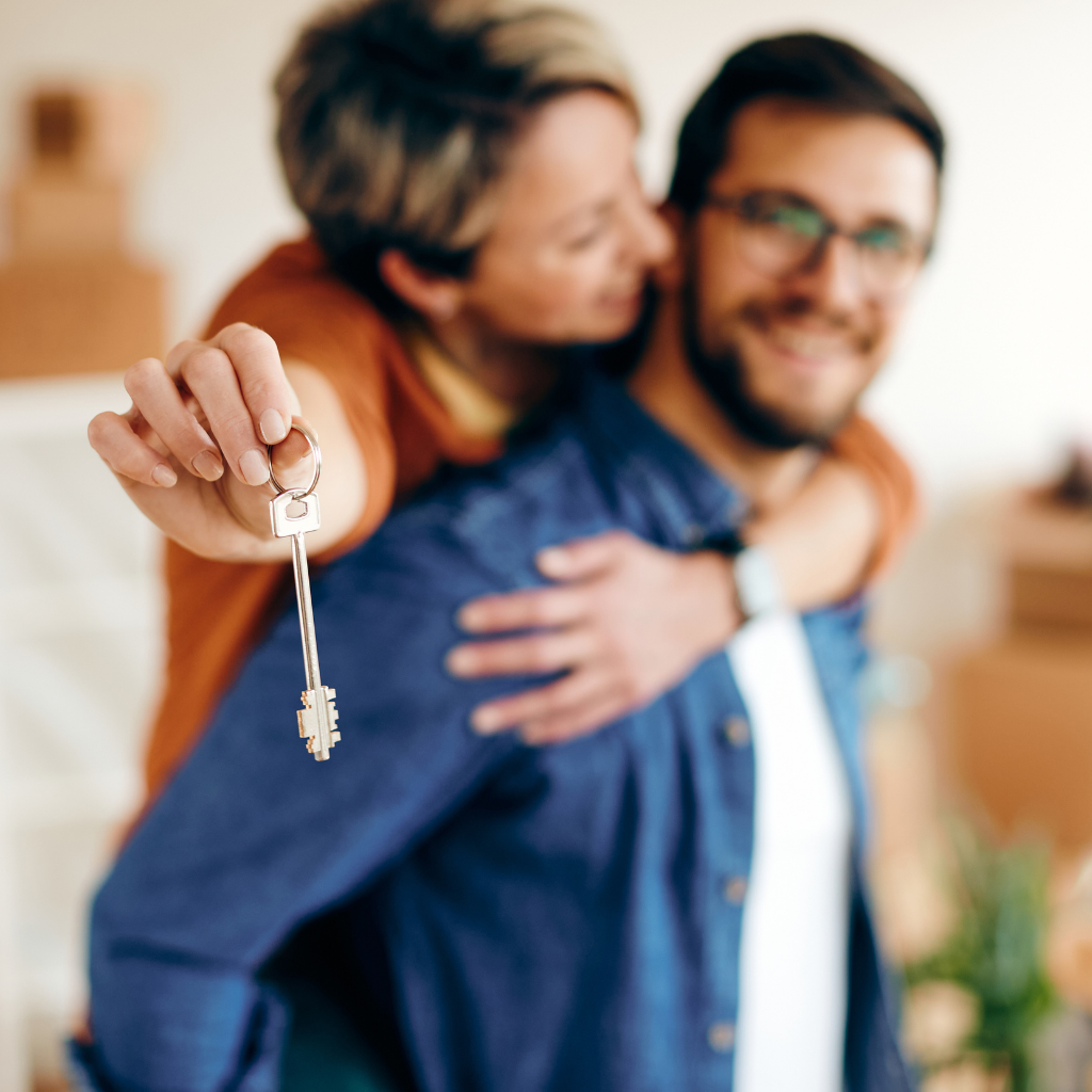 Woman holding keys, embracing smiling man; moving boxes in background.