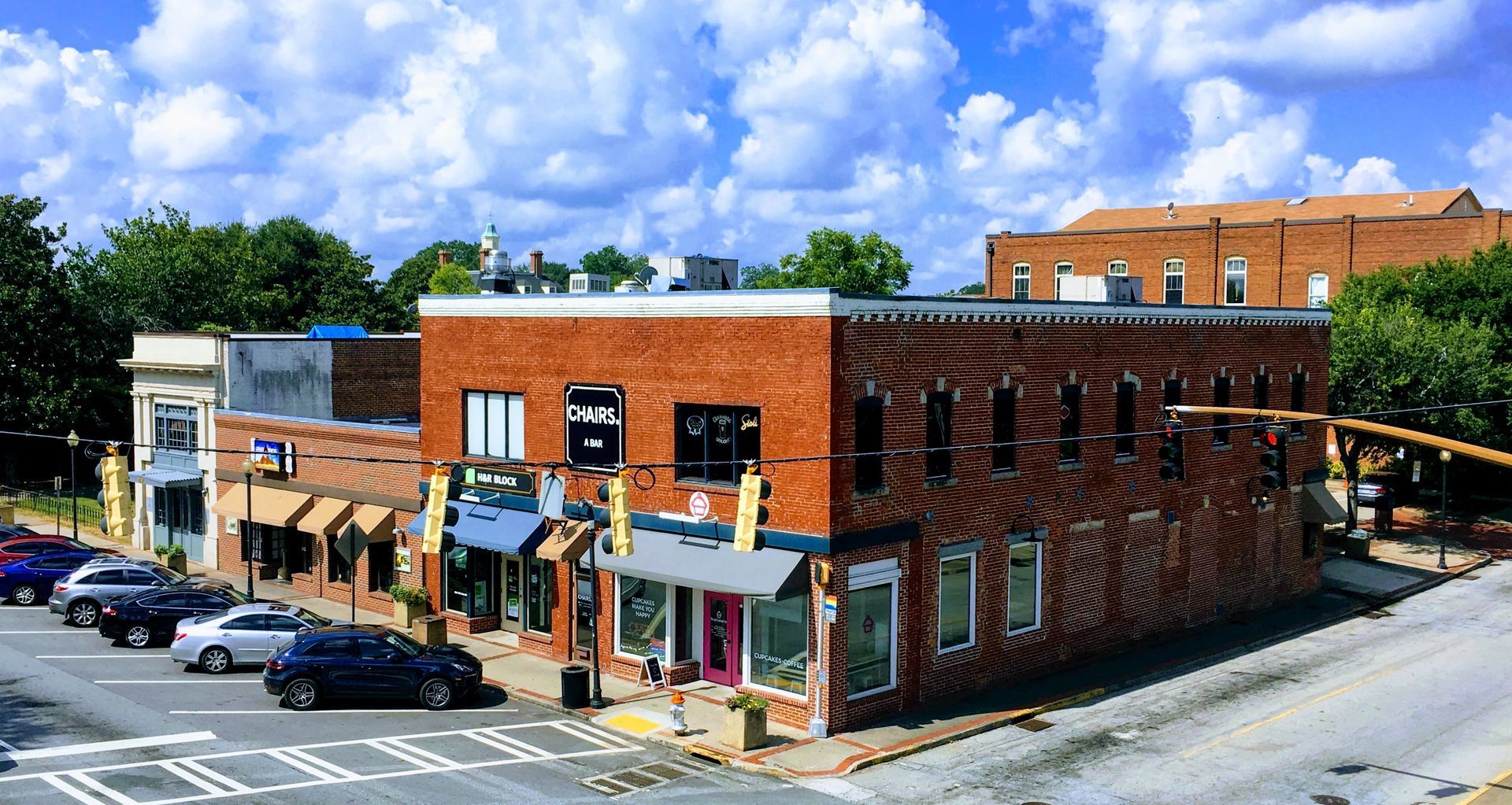 Buildings on a street corner, with parked cars. Brick facade and awnings, blue sky with clouds.