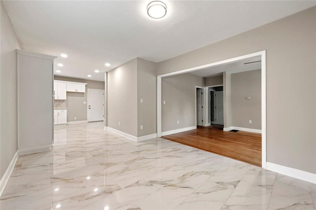 Empty, bright living room with marble floors and an open doorway to another room with wood flooring.