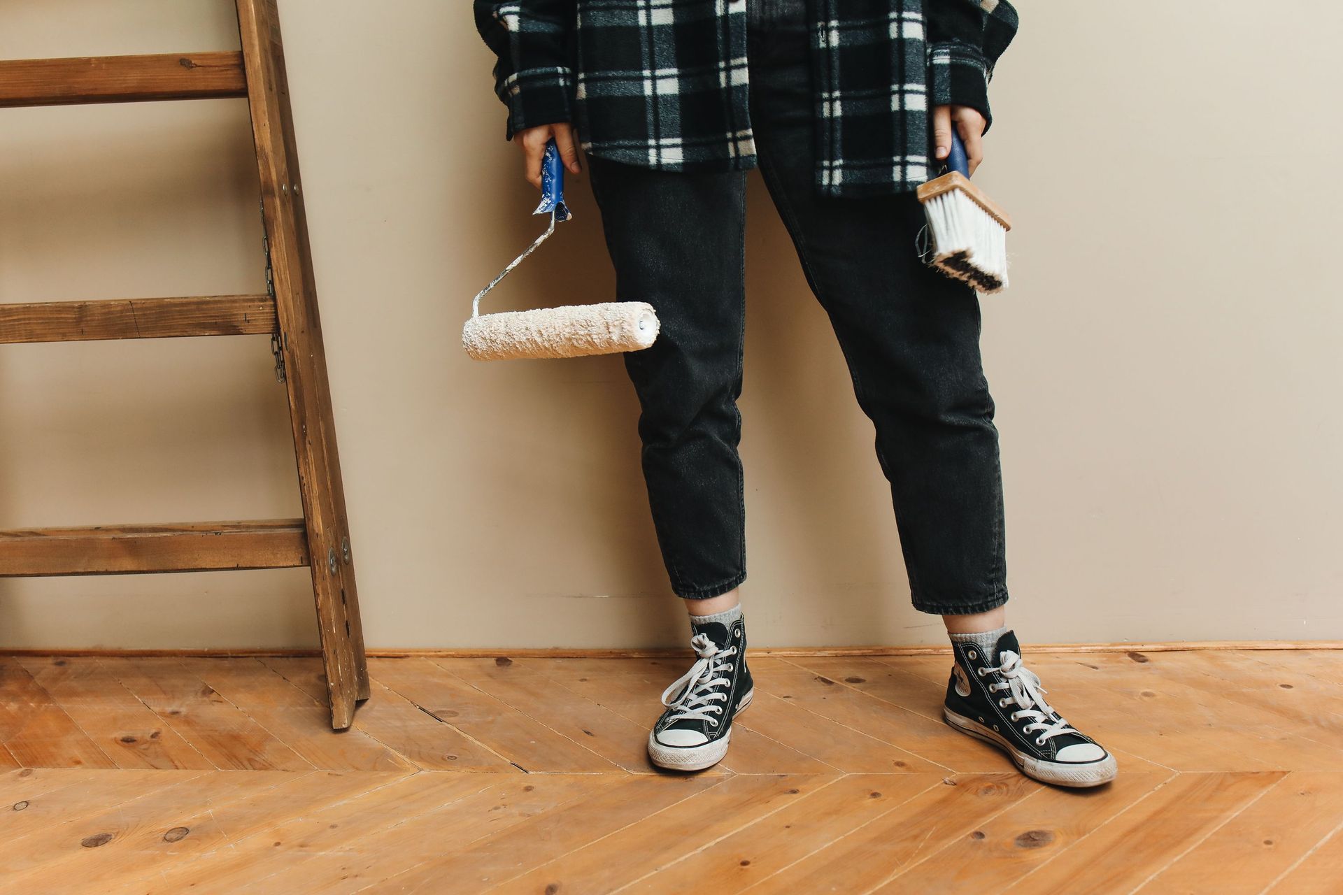 Person in black pants and sneakers standing indoors beside a wooden ladder, holding small items.