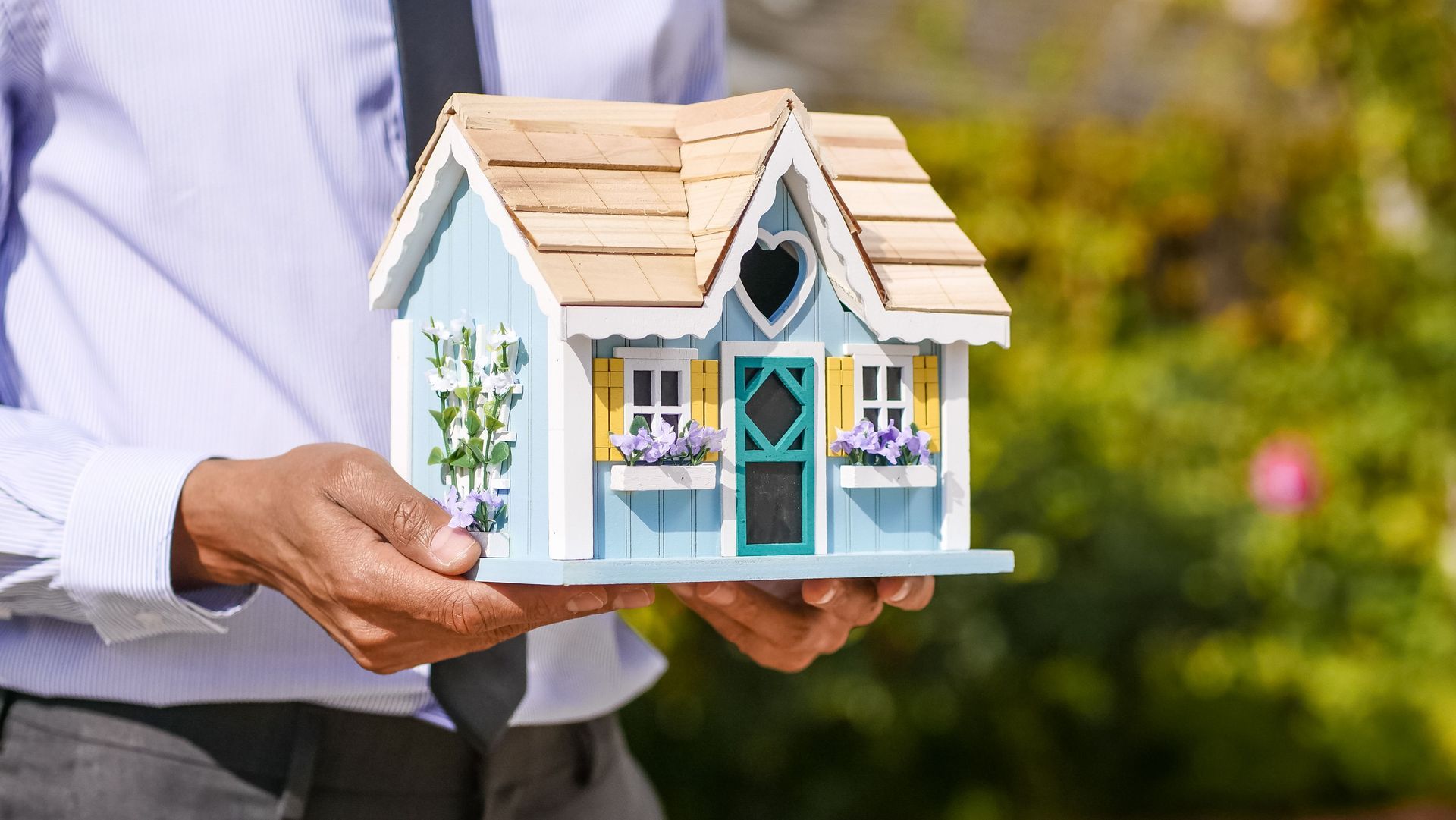 Hands holding a small pastel blue-and-white birdhouse model outdoors.