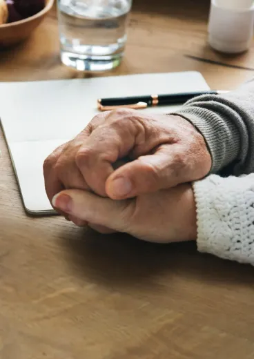 Two hands clasped on a wooden table beside a notebook and pencil.