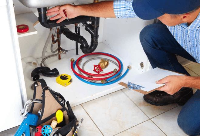 Plumber repairing sink pipes under a bathroom vanity with tools, red and blue hoses, and a clipboard nearby