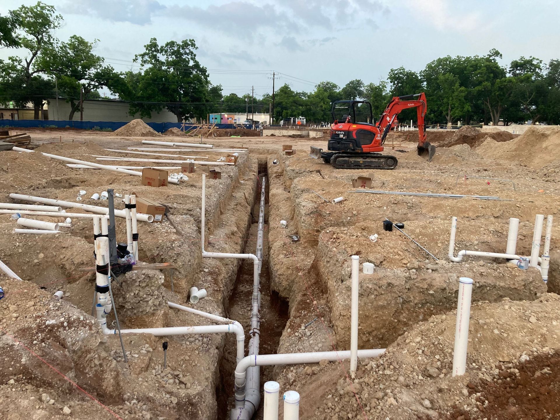 Construction site with an excavator digging a trench beside exposed white pipes and muddy ground
