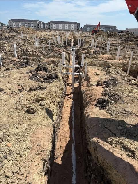 Construction site with trenches and wooden stakes, excavator in the background, and row houses beyond.