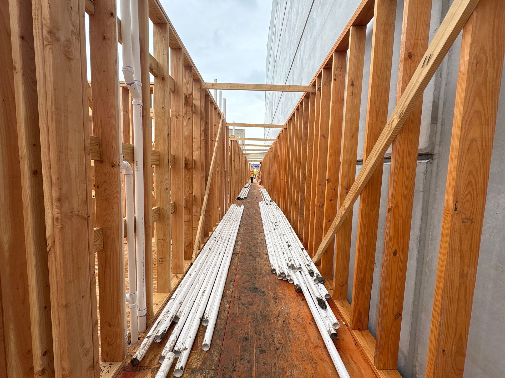 Wooden building frame with stacked white pipes along a dirt corridor under construction