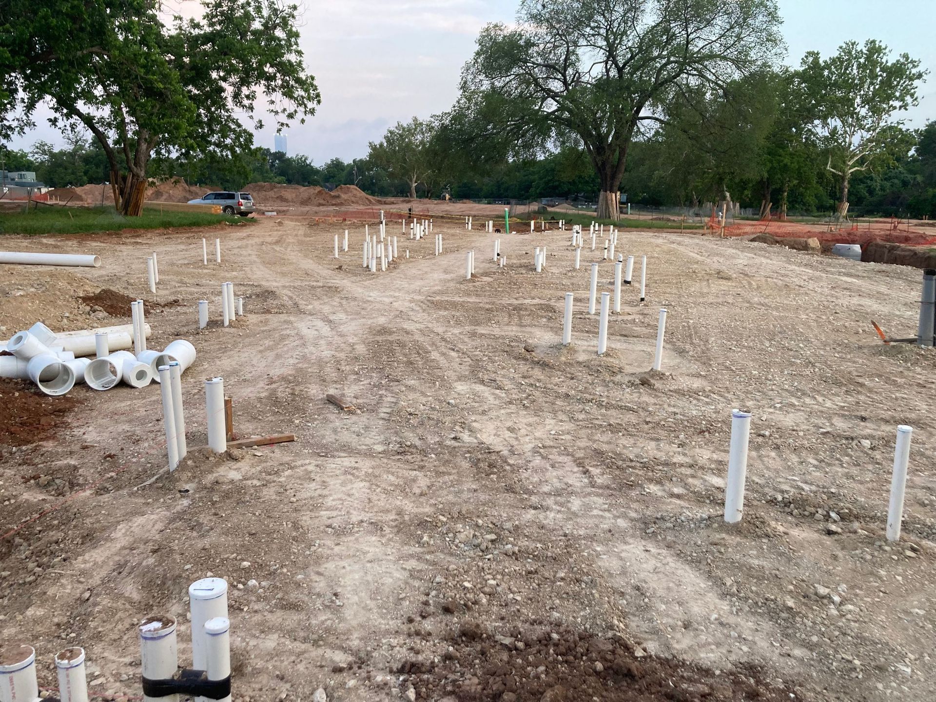 Construction site with white PVC pipes and markers scattered across a graded dirt lot under trees