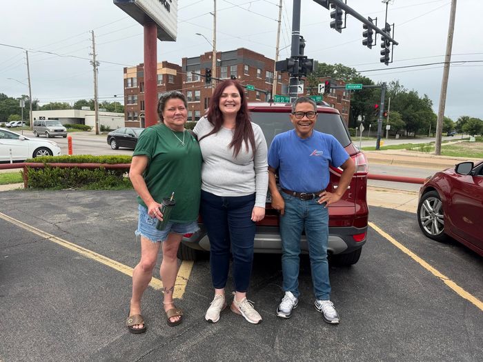 Three people posing in a parking lot beside a red car, with traffic lights and buildings behind them | Alexander’s Automotive, Inc.
