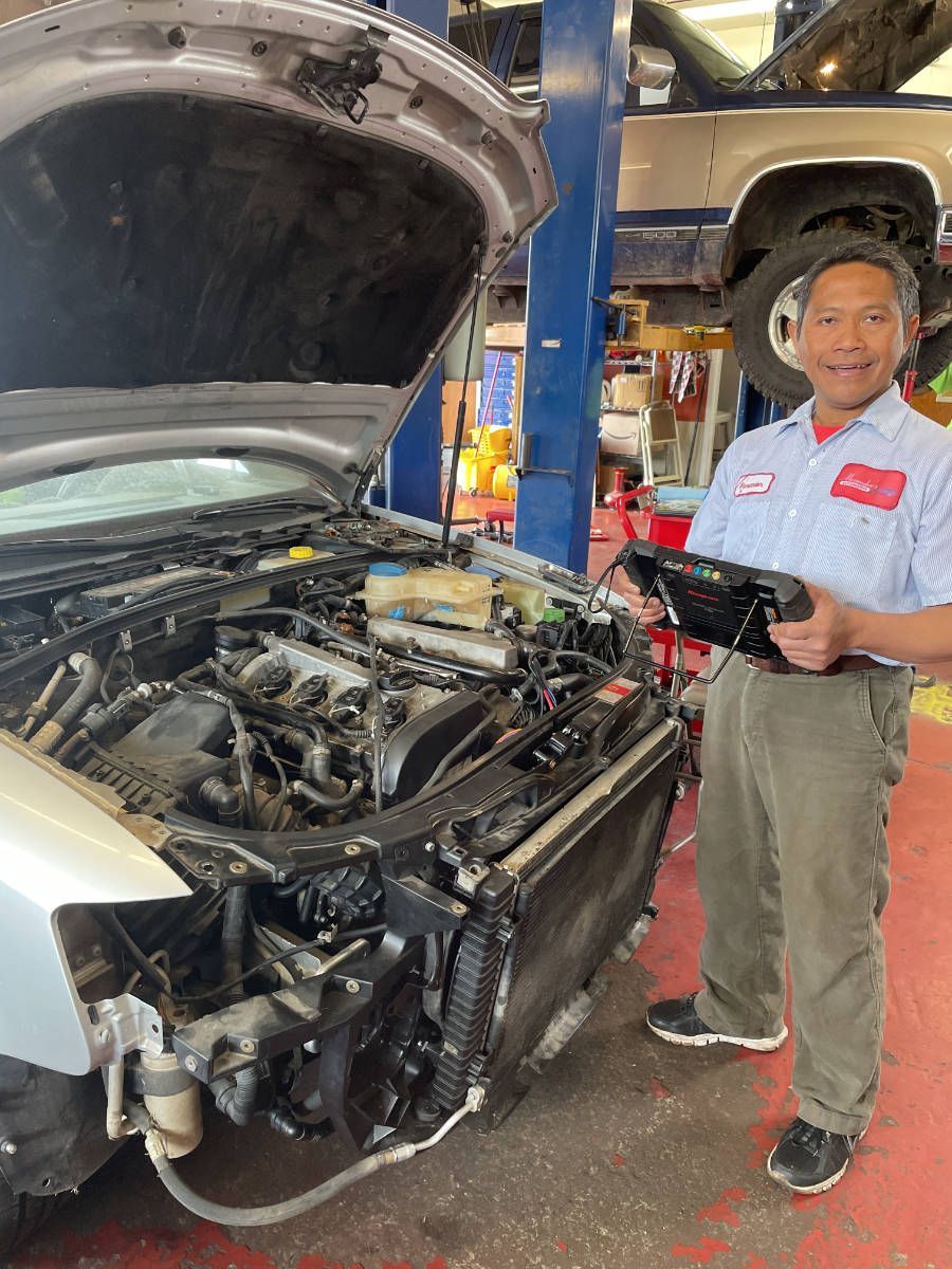 Mechanic standing beside a car with its hood open in an auto repair shop, holding tools | Alexander’s Automotive, Inc.