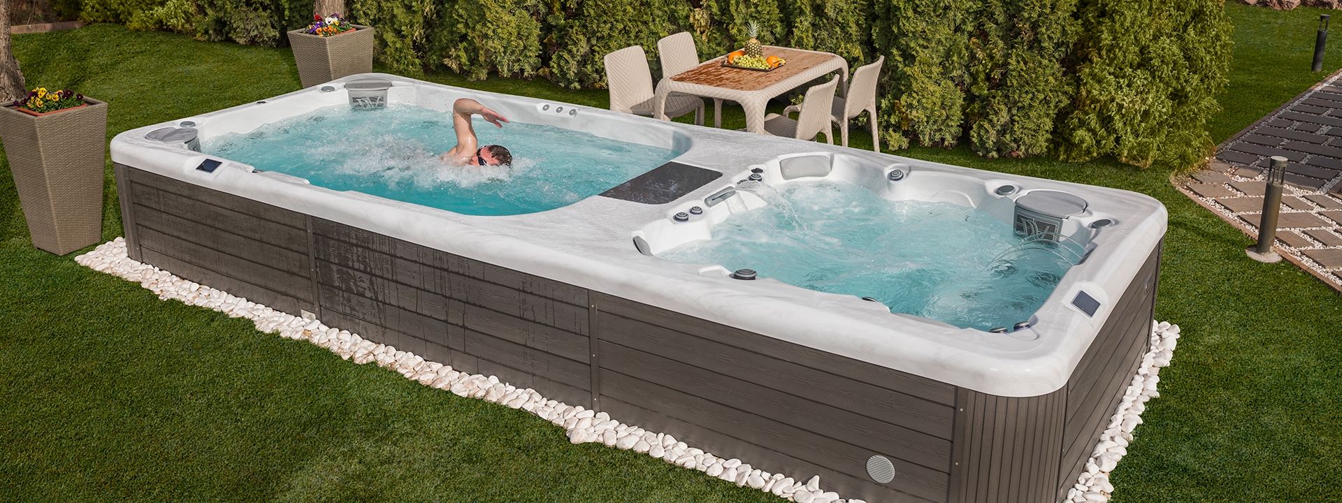 A swim spa with a person swimming. The spa is surrounded by grass and a hedge, with a table and chairs in the background.