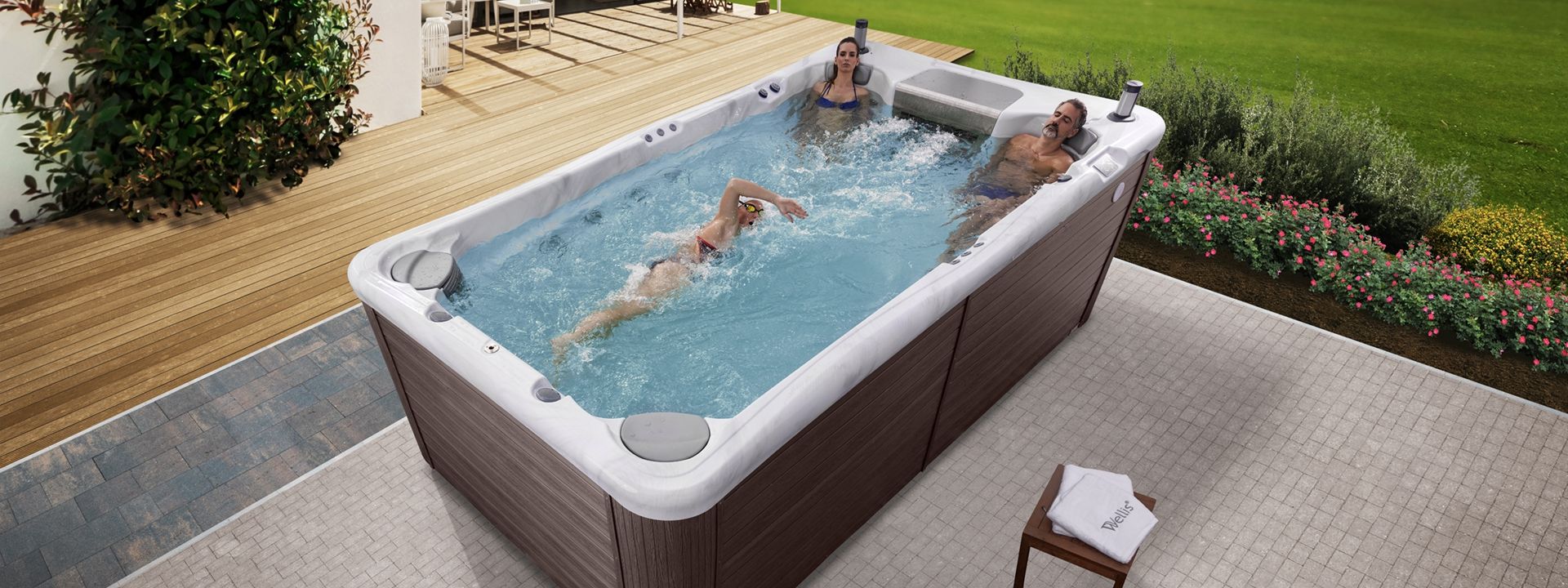 People in a swim spa on a patio. One person swims while others relax in the jets. Green lawn and deck nearby.