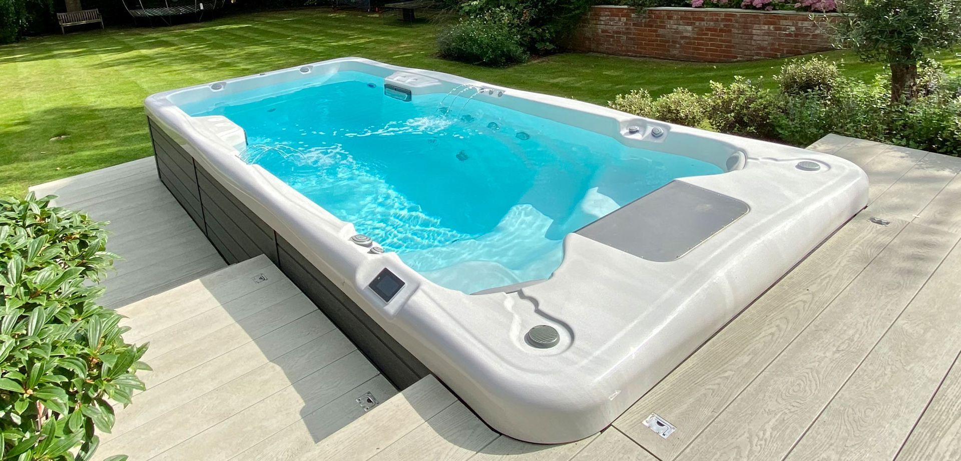 A rectangular, light-gray swim spa filled with blue water is in a backyard.