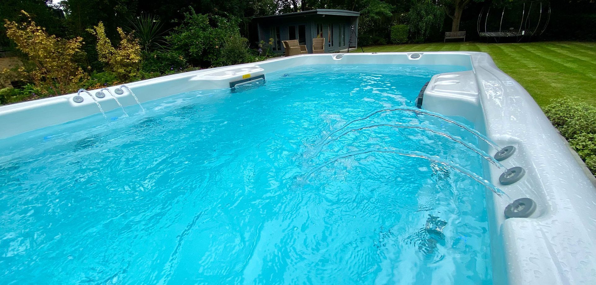 A rectangular, bright blue pool with water jets. A green lawn and trees surround the pool.