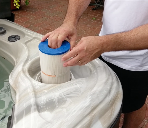 Person replacing a spa filter in a hot tub; hands holding a cylindrical blue-topped filter.