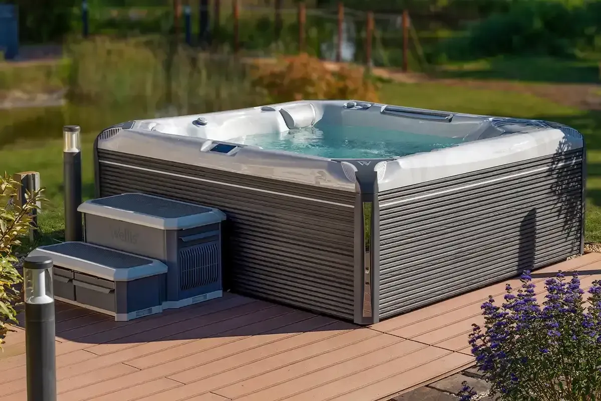 Hot tub on a wooden deck with steps. Water visible. Outdoor setting with greenery and lights.