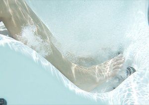 Person's legs underwater in a white hot tub, with bubbles and light reflections.