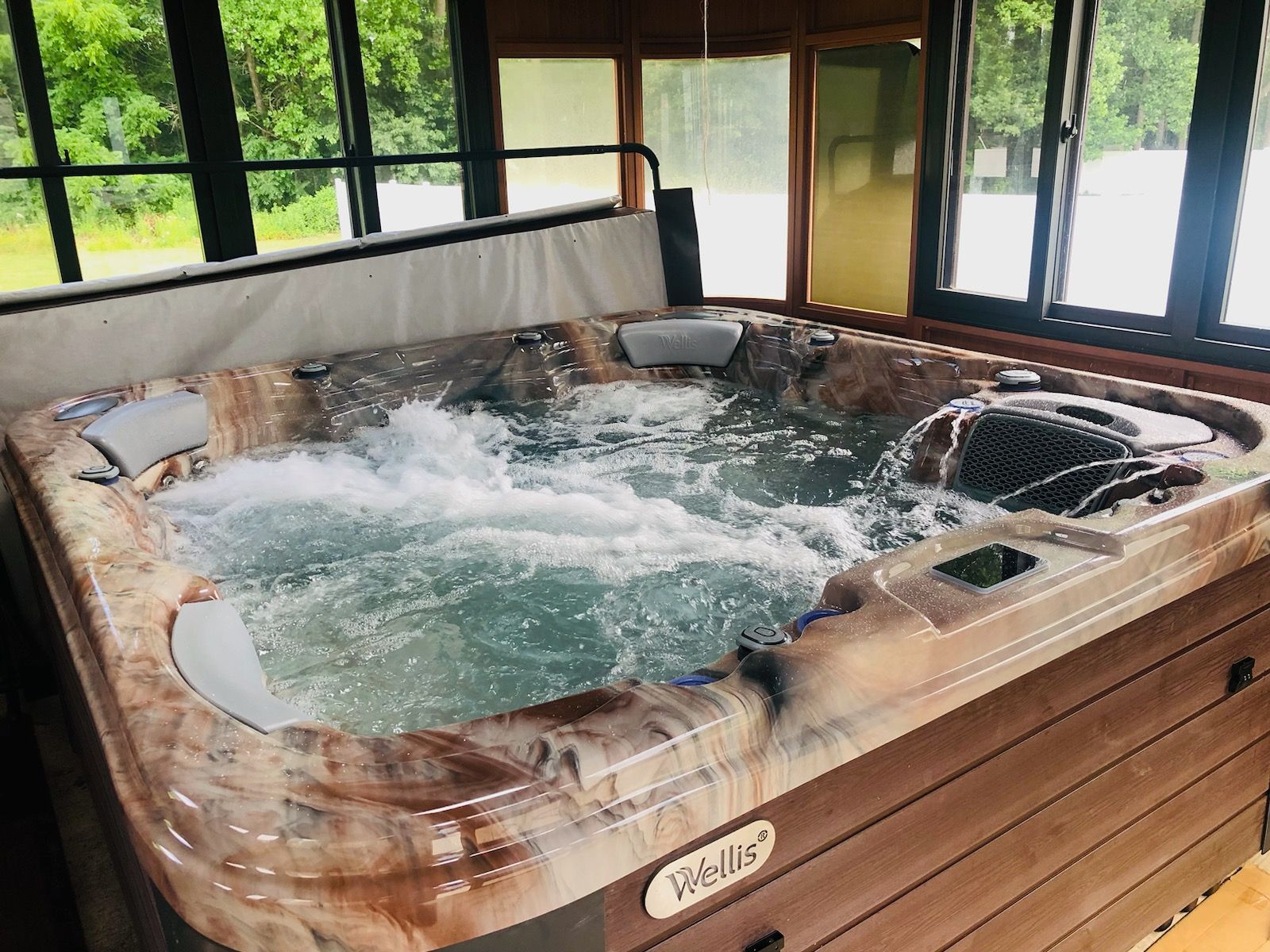 Hot tub with bubbling water in an enclosed room. Brown and gray tones, surrounded by windows with outdoor view.
