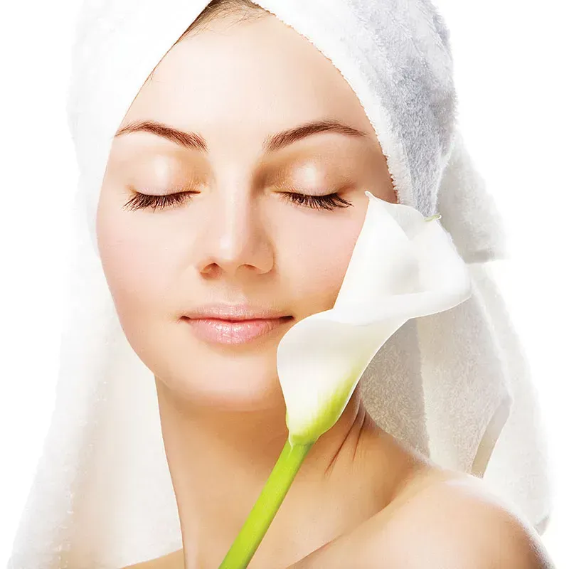 Woman with eyes closed, towel on head, holding white calla lily flower against cheek.