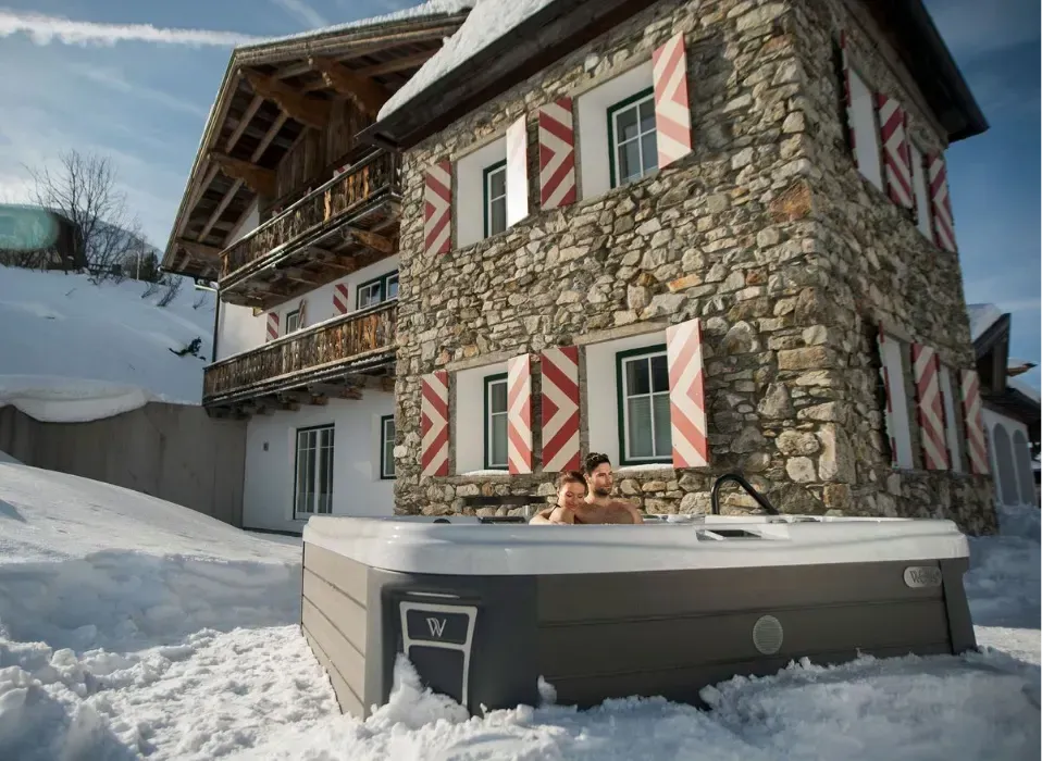 Two people in a hot tub outside a stone building with snow-covered surroundings.
