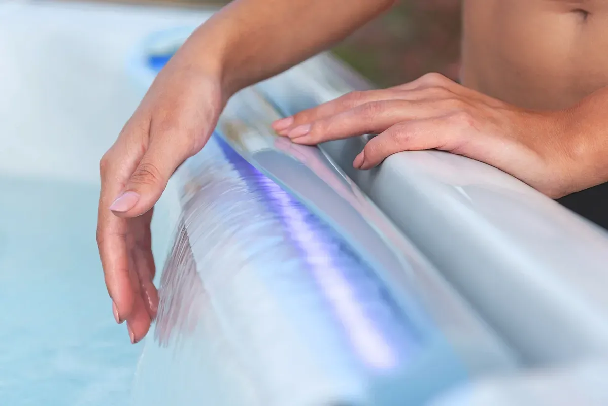 Person's hands resting on the edge of a hot tub with blue lights shining through the water.