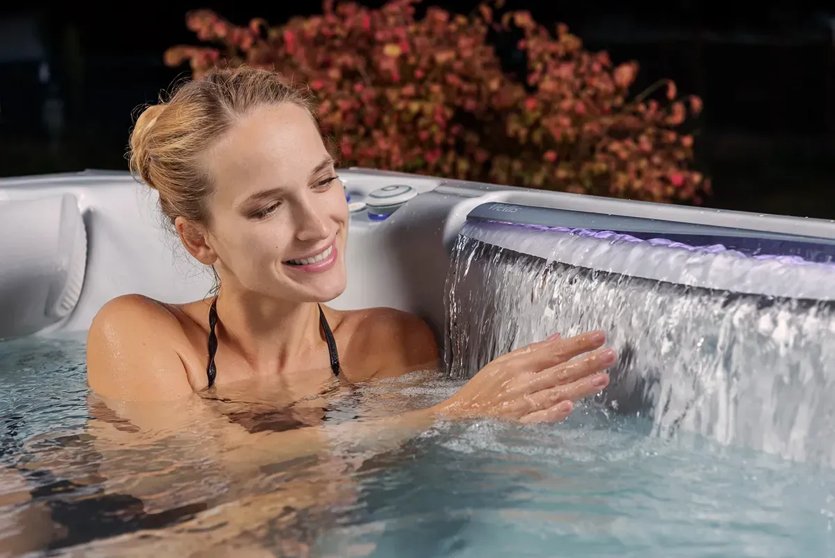 Woman smiling in a hot tub, touching a waterfall feature, night setting.