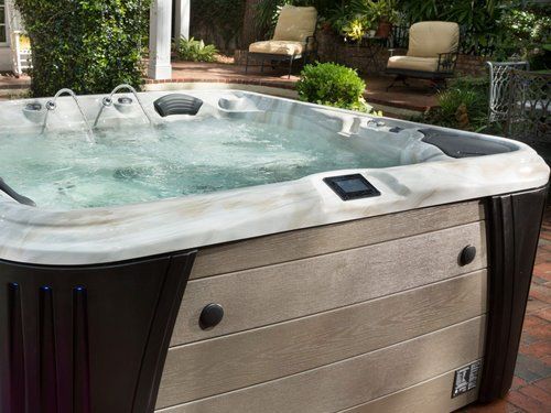 Hot tub with bubbling water, brown and tan exterior, set on brick patio with patio furniture in the background.