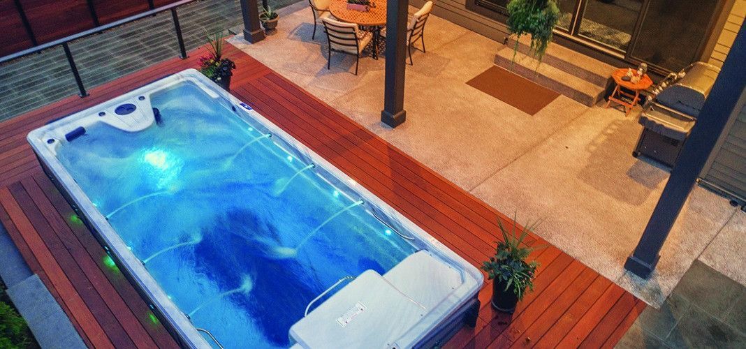 Outdoor hot tub on a wooden deck, with a seating area and greenery visible nearby.