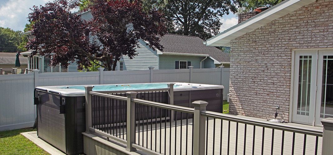 Backyard with a hot tub, deck, and a brick house. Green lawn and a white fence are also present.