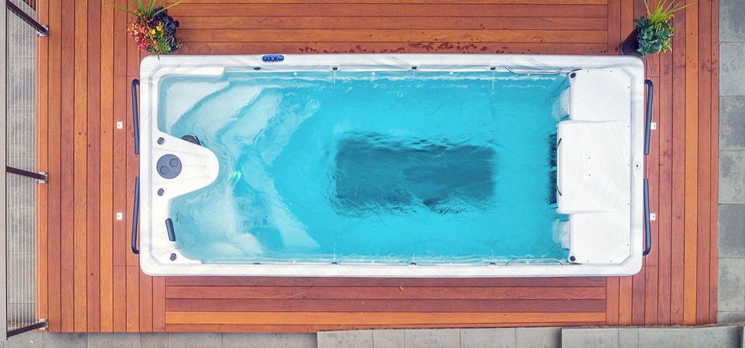 Overhead view of a rectangular, light-blue pool surrounded by wooden decking.  Green plants are at both ends of the pool.