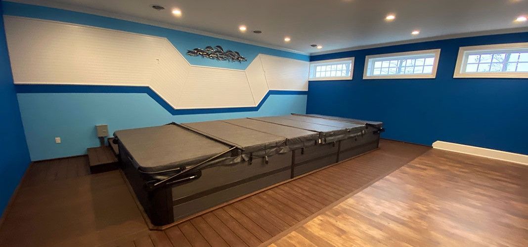 A large indoor swim spa in a room with blue walls, wood floors, and recessed lighting.