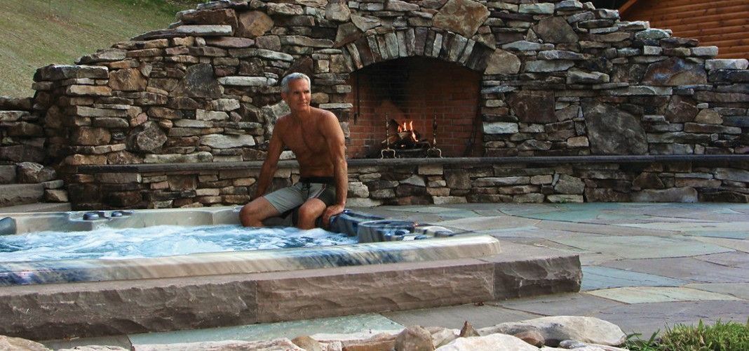 Man sits near hot tub, stone fireplace in the background, fire burning.