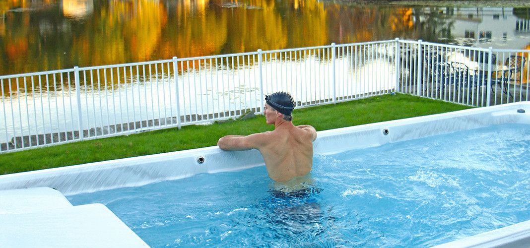 A man wearing a swim cap in an outdoor pool, overlooking a lake with trees.