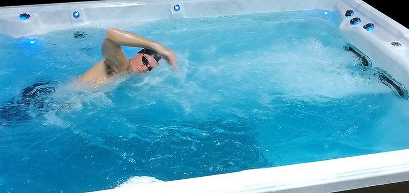 A man swims in a blue, rectangular pool with jets. He wears goggles and is in the water.