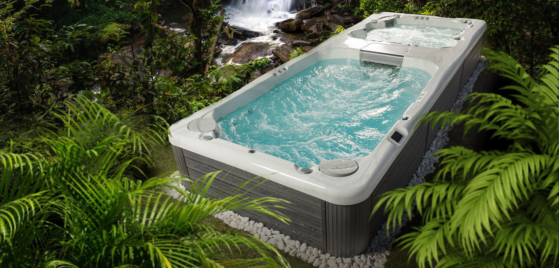 A rectangular swim spa surrounded by lush green foliage and a waterfall in the background.