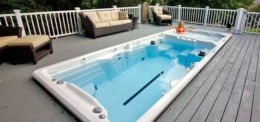 A long pool on a wooden deck with a person in the water, surrounded by seating and a railing.