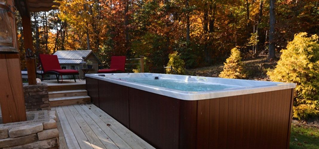 A rectangular hot tub surrounded by a wooden deck, set against a backdrop of autumn trees.