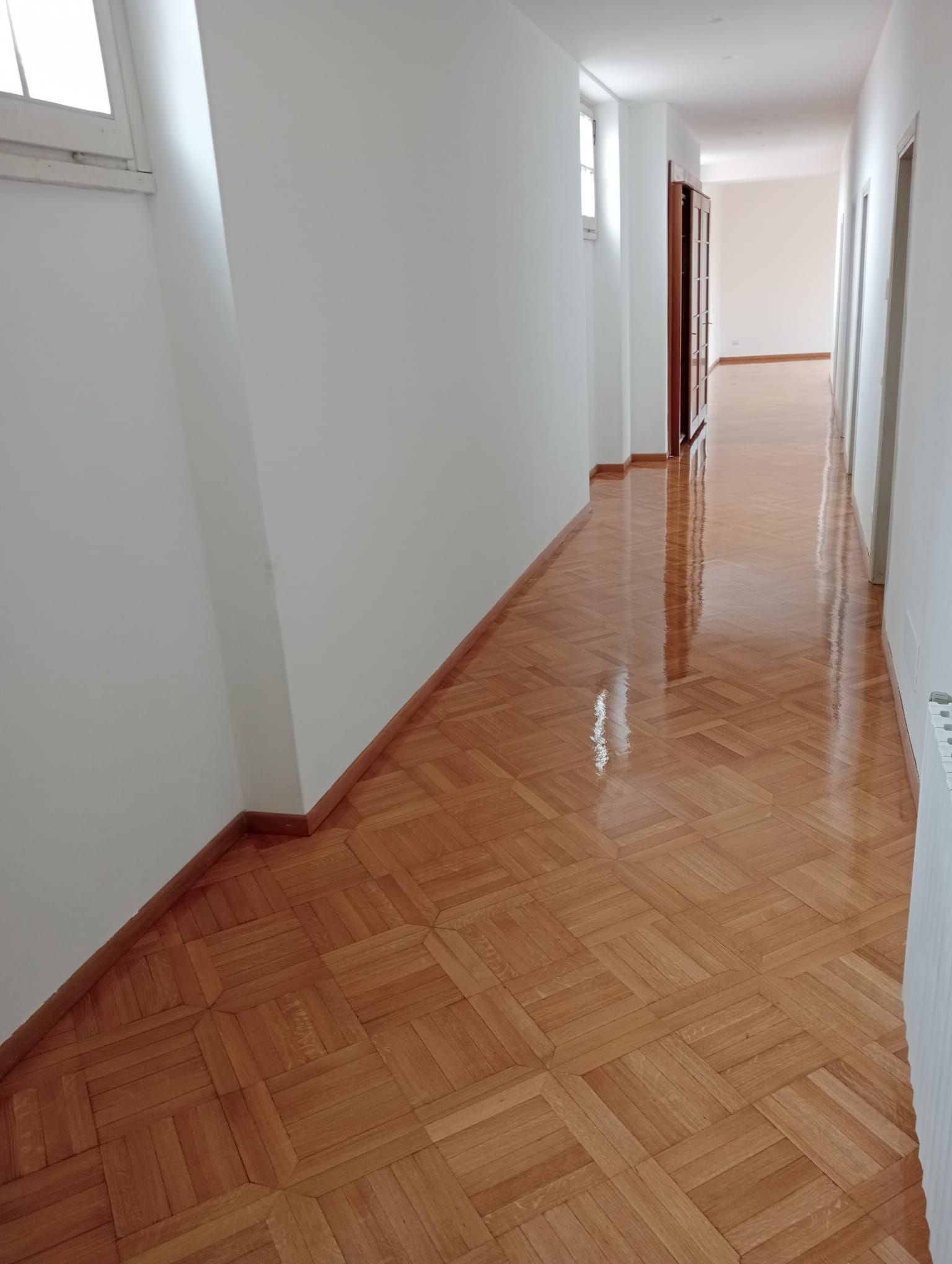 Corridoio lungo e vuoto, con pavimento in parquet chiaro e pareti bianche, illuminato dalle finestre.