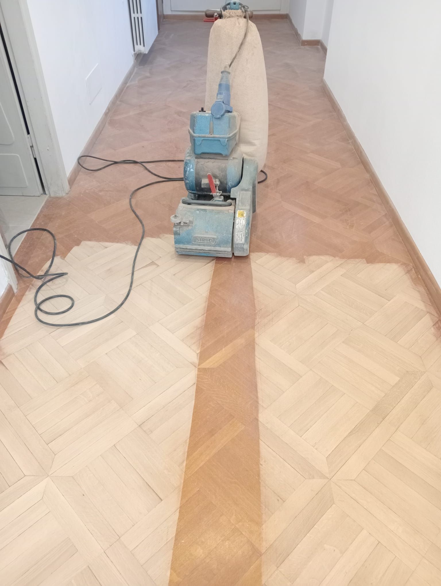 Per rifinire il pavimento in parquet di un corridoio, si utilizza una levigatrice per pavimenti, lasciando visibile una linea di legno levigato.