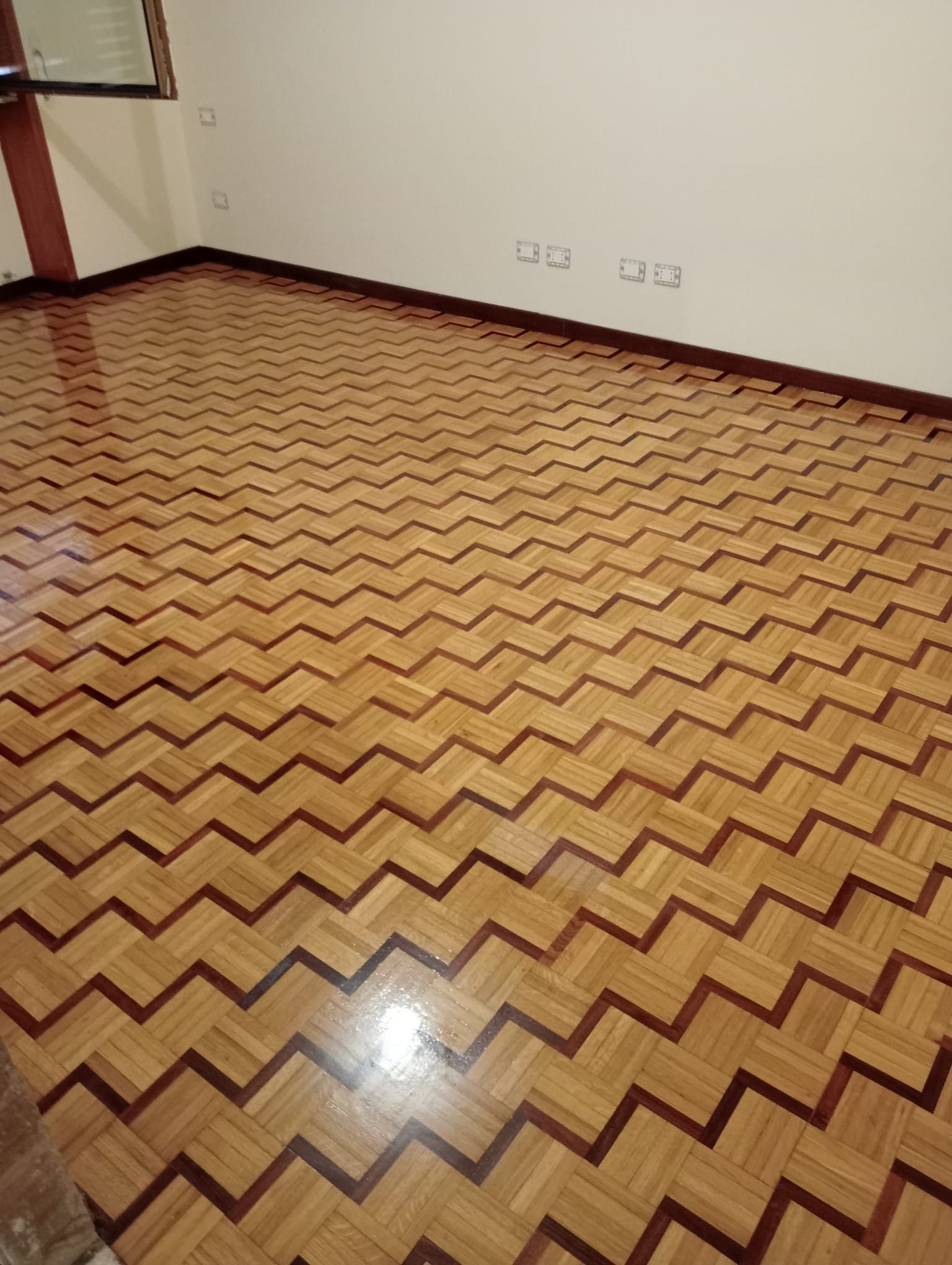 Pavimento in parquet con motivo a zigzag in una stanza con pareti bianche e bordo marrone.