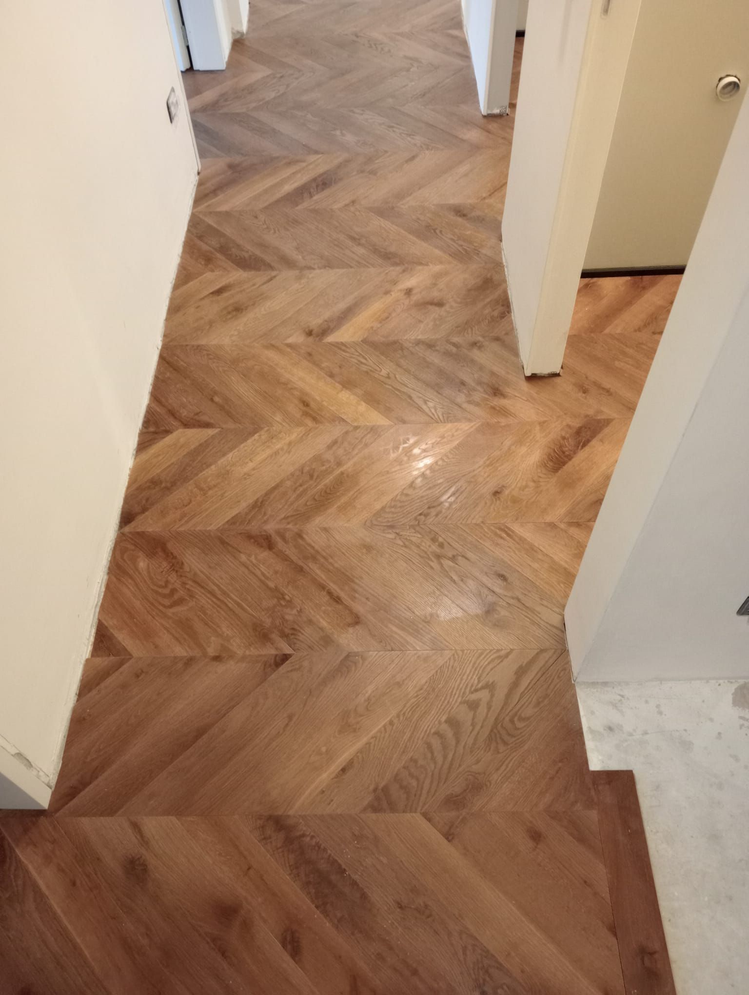 Pavimento in legno a spina di pesce in un corridoio, con pareti e porte di colore chiaro.
