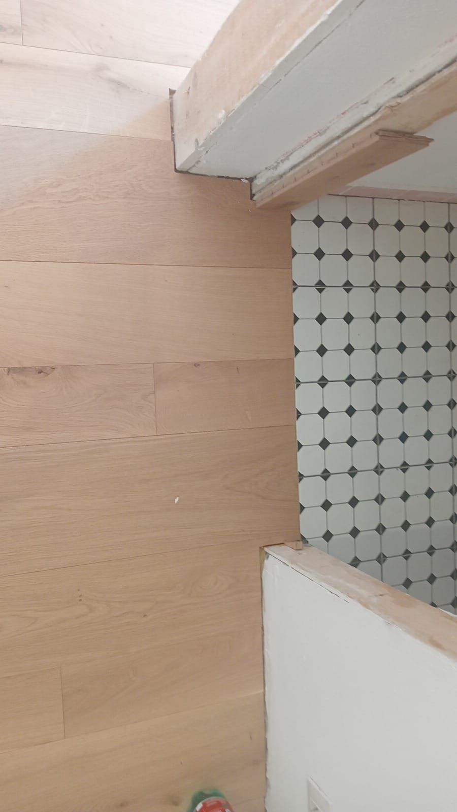 Un pavimento in legno chiaro incontra una zona piastrellata. La sezione piastrellata presenta un motivo a piccoli quadrati neri su sfondo bianco.