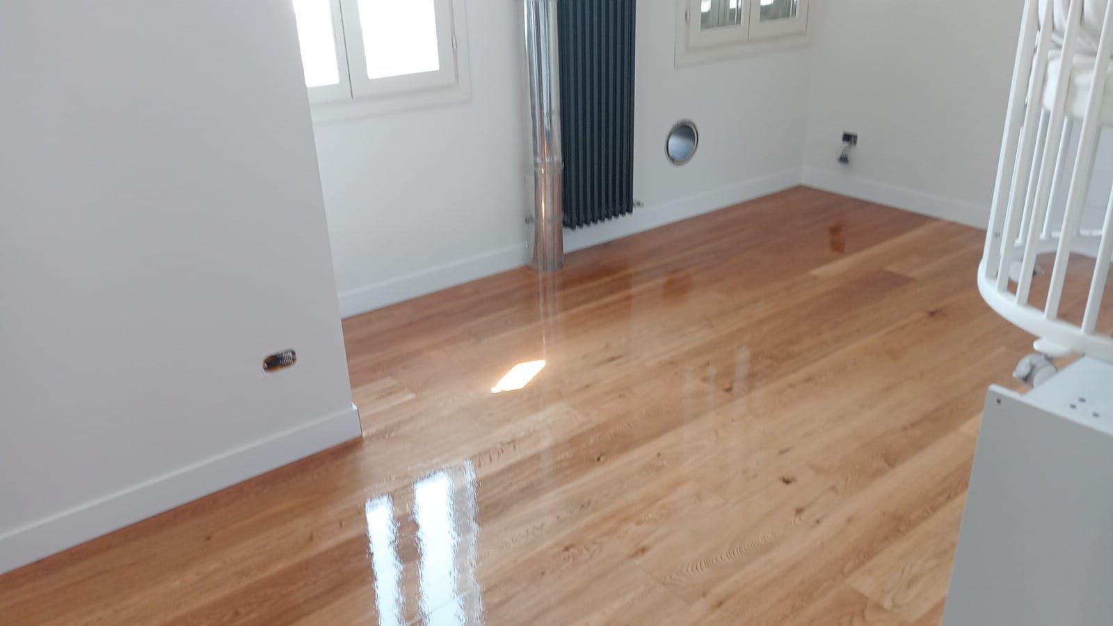 Pavimento in legno in una stanza con pareti bianche, finestre e un termosifone. La luce intensa si riflette sul legno lucido.
