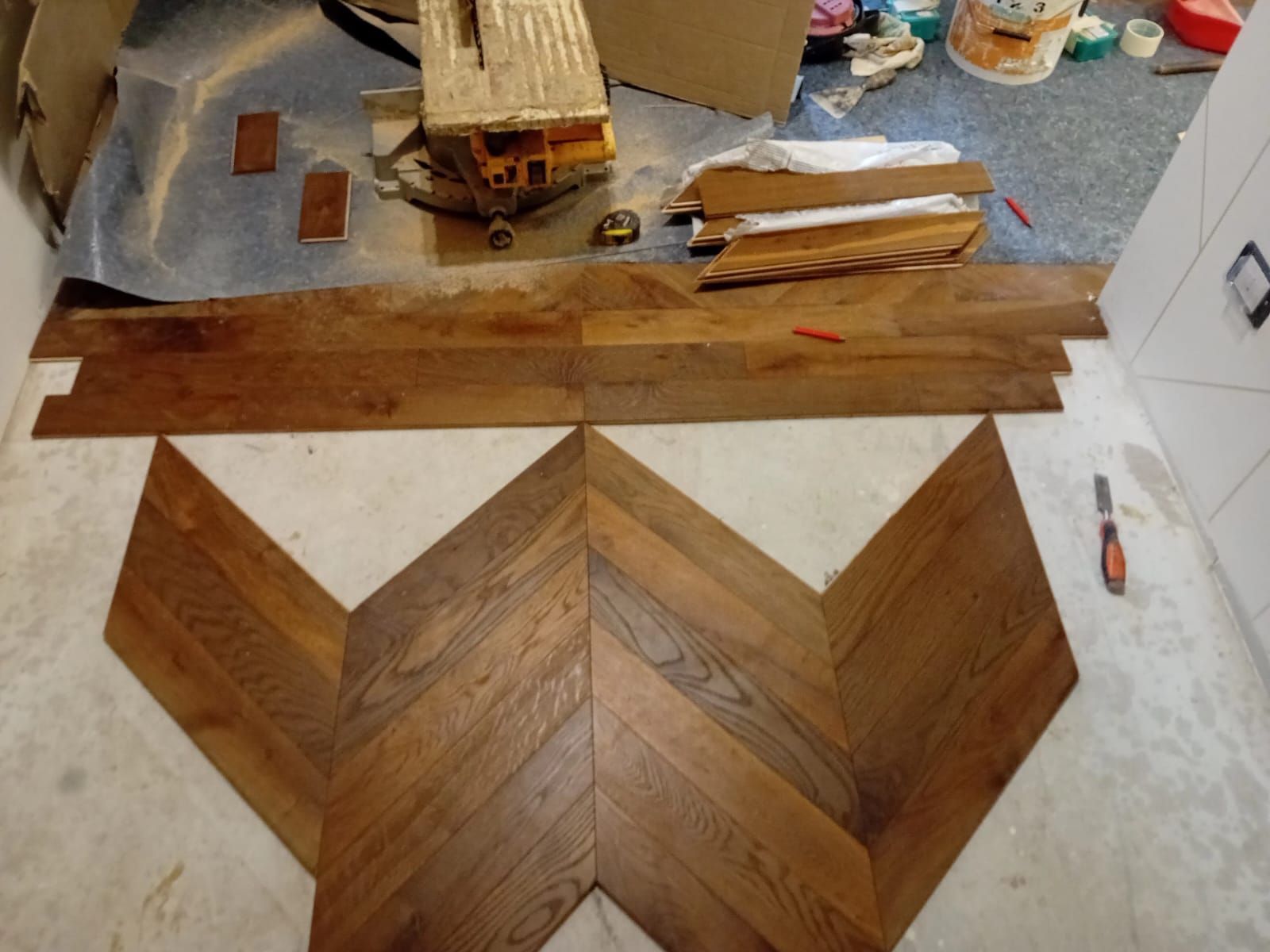 Pavimento in legno a spina di pesce in fase di installazione in una stanza. Sul pavimento sono disposti pezzi di legno marrone, alcuni dei quali ancora ammucchiati.