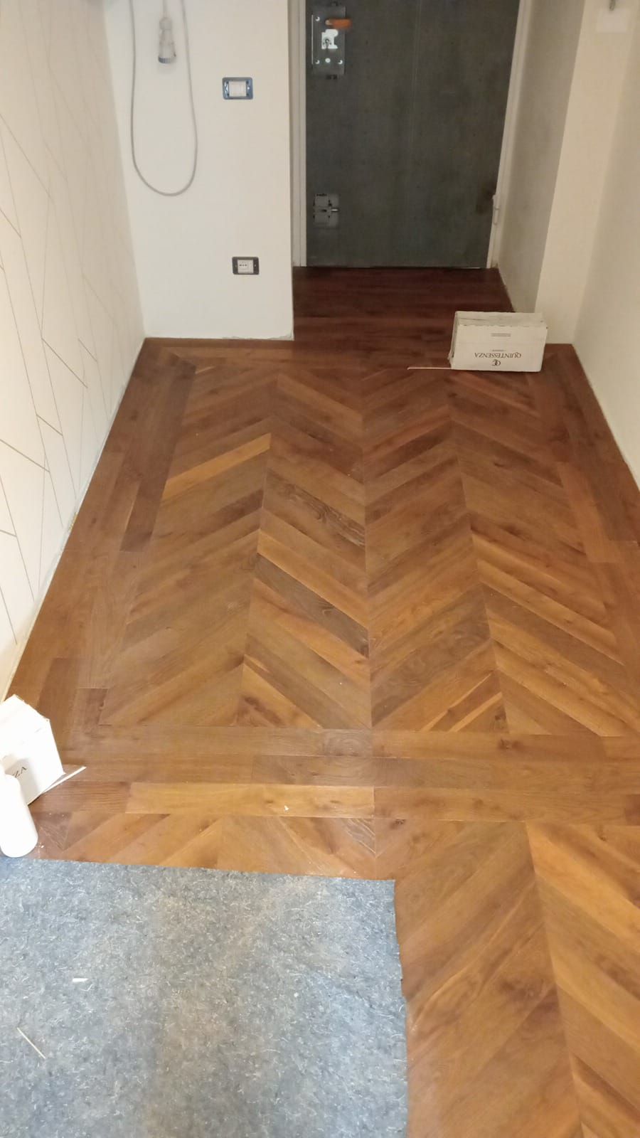 Pavimento in legno a spina di pesce in un corridoio con scale e porta. Il pavimento è di un caldo colore marrone.