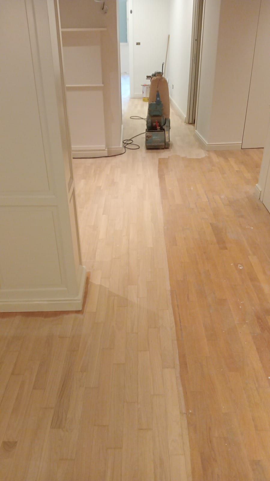 Corridoio con pavimento in legno grezzo in fase di levigatura, che mostra un netto contrasto tra le aree levigate e quelle non levigate. È visibile una levigatrice.