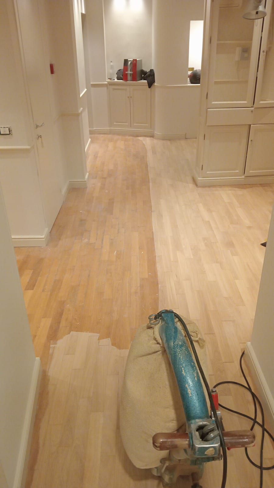 Un corridoio con pavimento in legno in fase di levigatura. La metà sinistra è grezza, con un colore legno chiaro, mentre la metà destra è levigata.