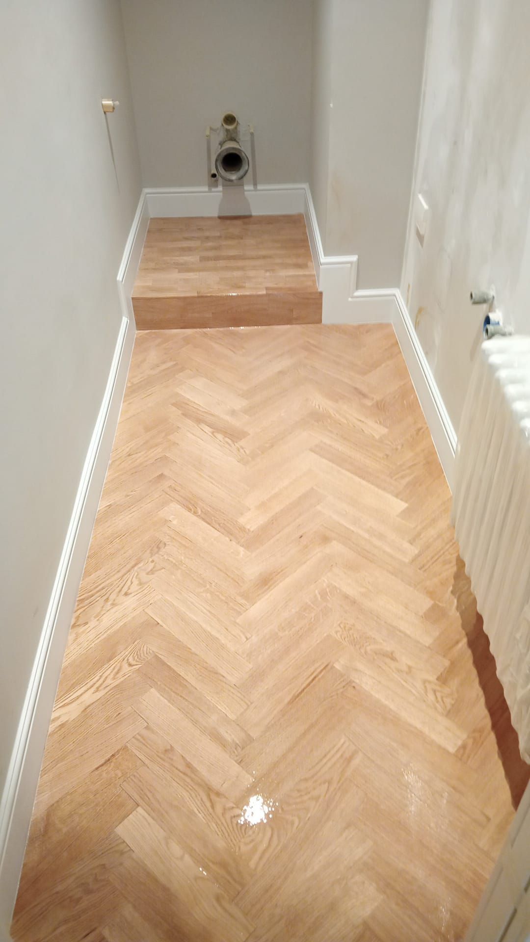 Pavimento in legno a spina di pesce che conduce a una piccola piattaforma rialzata con WC. Rifiniture e pareti bianche.