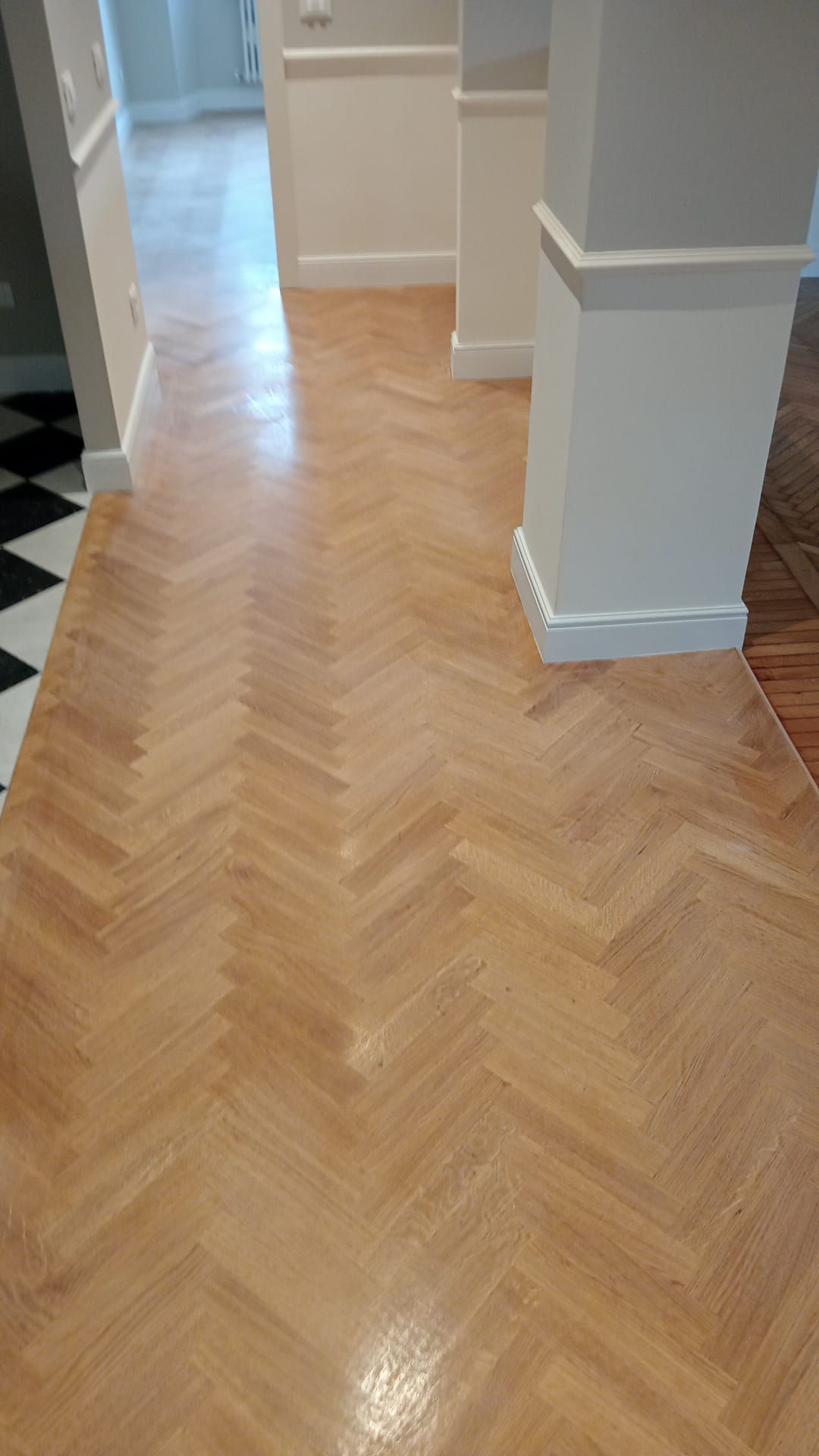Pavimento in legno chiaro con motivo a spina di pesce e finitura lucida, pareti quasi bianche e una colonna.